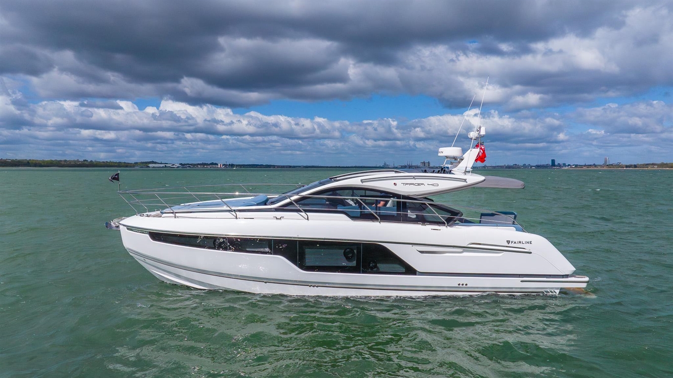 A sleek white Fairline Targa 40 yacht glides across turquoise waters under a cloudy sky, seen from a side angle.
